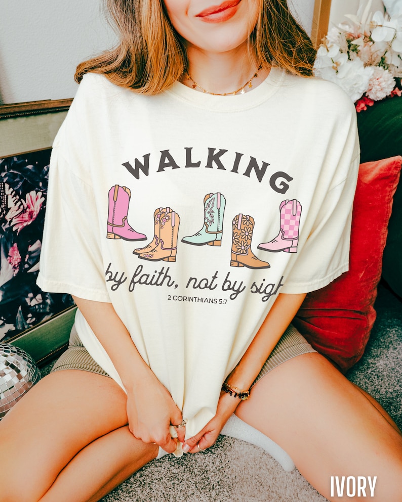 Puede incluir: Camiseta de color marfil con el texto "WALKING by faith, not by sight" y una fila de cinco botas de vaquero de colores. El texto "2 CORINTHIANS 5:7" est&aacute; debajo de las botas.