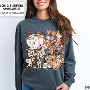 Puede incluir: Una camiseta de manga larga gris oscuro con un dise&ntilde;o de tem&aacute;tica oto&ntilde;al. El dise&ntilde;o incluye calabazas florales, lazos y las palabras "Autumn Vibes", "Cozy" y "Harvest". La camiseta tiene el texto "Long Sleeves Available Choose @ Checkout."