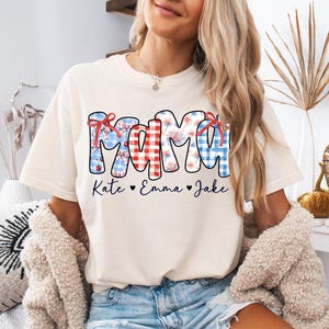 May include: Ivory-colored t-shirt with the word "MAMA" in a patriotic design. The letters are decorated with red, white, and blue patterns, including gingham and fireworks. Below the word are the names "Kate," "Emma," and "Jake."