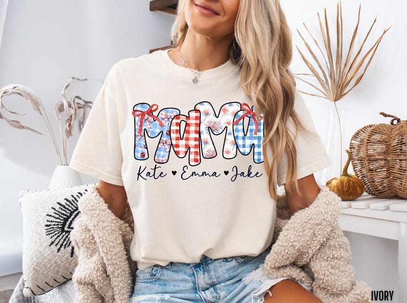 May include: Ivory-colored t-shirt with the word "MAMA" in a patriotic design. The letters are decorated with red, white, and blue patterns, including gingham and fireworks. Below the word are the names "Kate," "Emma," and "Jake."