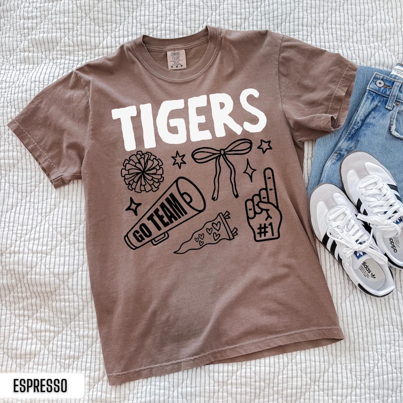 Puede incluir: Una camiseta marr&oacute;n de manga corta con el texto blanco TIGERS y gr&aacute;ficos de l&iacute;neas negras que incluyen un pomp&oacute;n, un lazo, un meg&aacute;fono con el texto GO TEAM, un bander&iacute;n con corazones y un dedo de espuma con el n&uacute;mero 1.