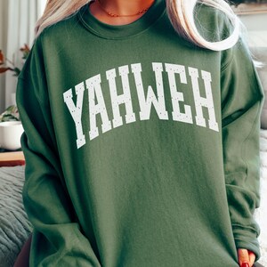 May include: A military green sweatshirt with the word "YAHWEH" in large, distressed white letters across the chest. The sweatshirt has a crew neck and long sleeves. The words "MILITARY GREEN" are printed at the bottom left.