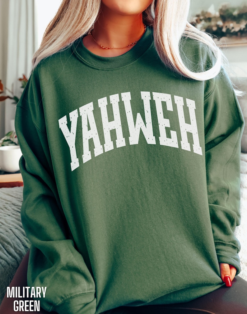 May include: A military green sweatshirt with the word "YAHWEH" in large, distressed white letters across the chest. The sweatshirt has a crew neck and long sleeves. The words "MILITARY GREEN" are printed at the bottom left.