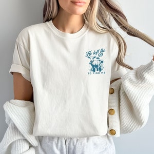 May include: Cream-coloured t-shirt with rolled sleeves. A teal graphic on the front features a cow and the text "He left the 99 to find me." The shirt is made of soft material.