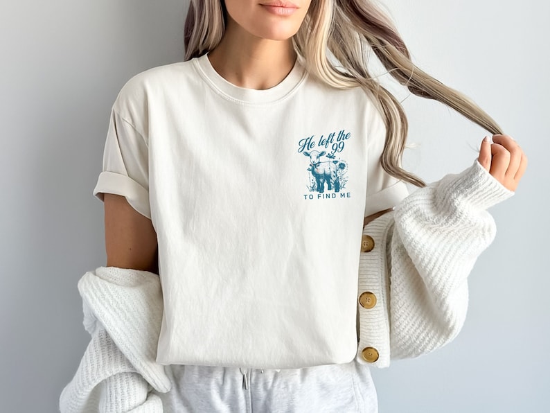 May include: Cream-coloured t-shirt with rolled sleeves. A teal graphic on the front features a cow and the text "He left the 99 to find me." The shirt is made of soft material.