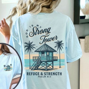 Puede incluir: Camiseta azul claro con un gr&aacute;fico de una escena de playa con una torre de salvavidas y palmeras. El texto "God is my Strong Tower" y "Refuge & Strength Psalm 61:3" est&aacute; impreso en la camiseta.