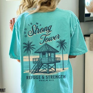 Puede incluir: Una camiseta azul turquesa con un gr&aacute;fico de una escena de playa con una torre de salvavidas y palmeras. El texto "God is my Strong Tower" est&aacute; escrito en una fuente cursiva sobre el gr&aacute;fico. El texto "Refuge & Strength Psalm 61:3" est&aacute; escrito debajo del gr&aacute;fico.