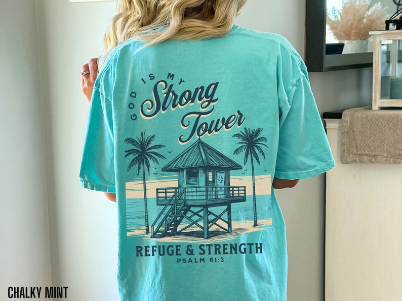 Puede incluir: Una camiseta azul turquesa con un gr&aacute;fico de una escena de playa con una torre de salvavidas y palmeras. El texto "God is my Strong Tower" est&aacute; escrito en una fuente cursiva sobre el gr&aacute;fico. El texto "Refuge & Strength Psalm 61:3" est&aacute; escrito debajo del gr&aacute;fico.