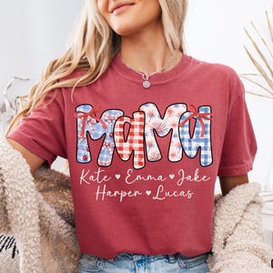 May include: Crimson-colored t-shirt with the word "MAMA" in a patriotic design, featuring red, white, and blue patterns. Below the word are the names Kate, Emma, Jake, Harper, and Lucas. The shirt is a comfortable, casual style.