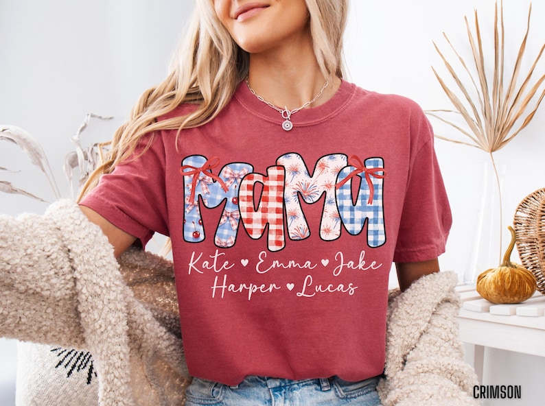 May include: Crimson-colored t-shirt with the word "MAMA" in a patriotic design, featuring red, white, and blue patterns. Below the word are the names Kate, Emma, Jake, Harper, and Lucas. The shirt is a comfortable, casual style.