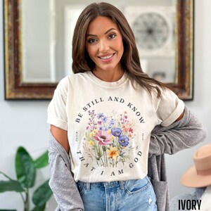 May include: A beige t-shirt with a floral design and the text "Be still and know that I am God" in a circle around the flowers.