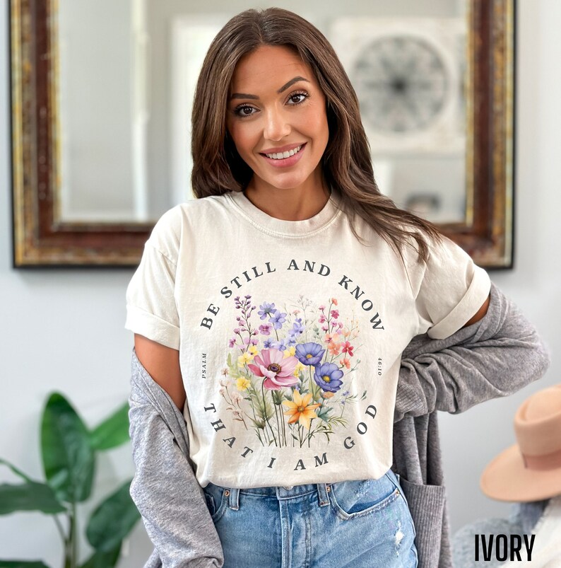 May include: A beige t-shirt with a floral design and the text "Be still and know that I am God" in a circle around the flowers.
