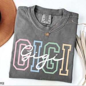 May include: Gray t-shirt with the word "Gigi" in white script lettering inside of colorful outlined block letters. The shirt has a tag that says "Comfort Colors" and "S - P - CH".