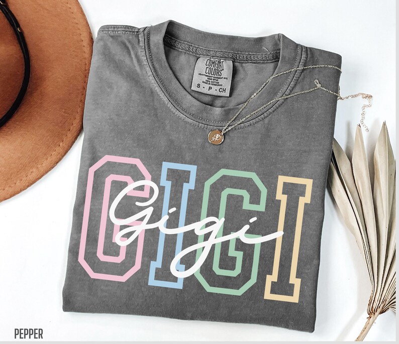 May include: Gray t-shirt with the word "Gigi" in white script lettering inside of colorful outlined block letters. The shirt has a tag that says "Comfort Colors" and "S - P - CH".