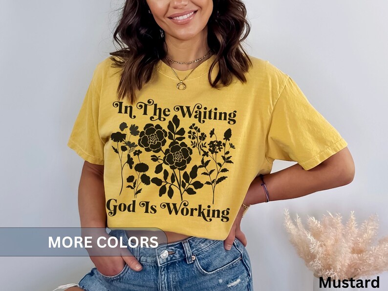 Op de afbeelding: Een mosterdgeel T-shirt met een zwart bloemenontwerp en de tekst "In the waiting God is working".