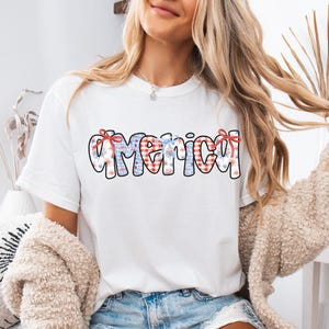 May include: White t-shirt with a red, white, and blue "America" graphic featuring a checkered pattern and fireworks. The graphic is centered on the shirt.