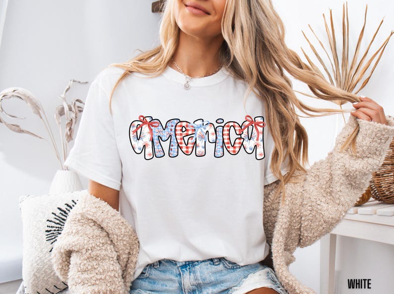 May include: White t-shirt with a red, white, and blue "America" graphic featuring a checkered pattern and fireworks. The graphic is centered on the shirt.