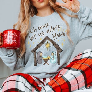 Puede incluir: Una sudadera gris con el texto "Oh come let us adore Him" y una escena de la natividad de dibujos animados. La sudadera la lleva puesta una persona que sostiene una taza roja con texto blanco que dice "Have yourself a merry little Christmas".