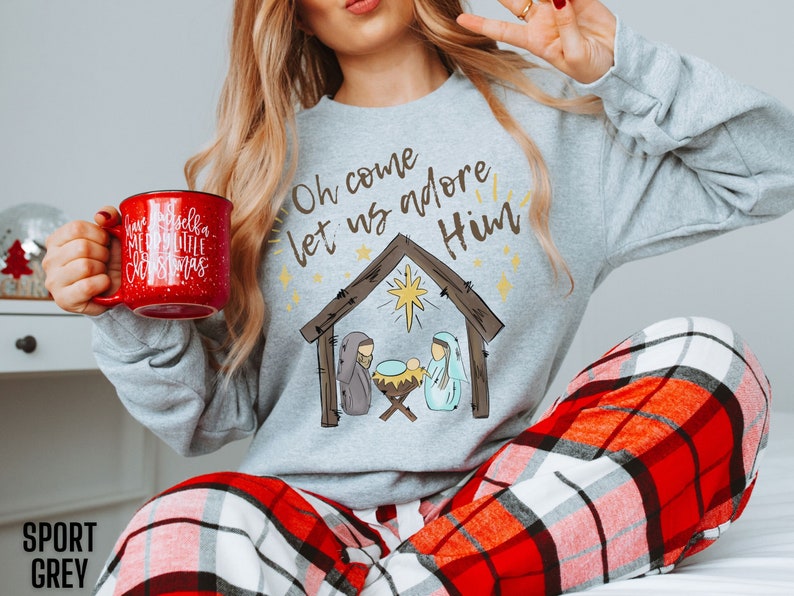 Puede incluir: Una sudadera gris con el texto "Oh come let us adore Him" y una escena de la natividad de dibujos animados. La sudadera la lleva puesta una persona que sostiene una taza roja con texto blanco que dice "Have yourself a merry little Christmas".