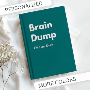 May include: A teal-colored journal with the words "Brain Dump" in large white letters. Below, the text reads "Of: Cam Smith." The word "PERSONALIZED" is above the journal, and "MORE COLORS" is below. The journal is on a white surface.