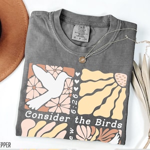 Op de afbeelding: Een grijs T-shirt met een grafisch ontwerp van een witte vogel, bloemen en de tekst "Consider the Birds 6:26".