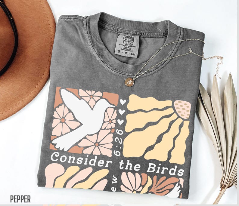 Op de afbeelding: Een grijs T-shirt met een grafisch ontwerp van een witte vogel, bloemen en de tekst "Consider the Birds 6:26".