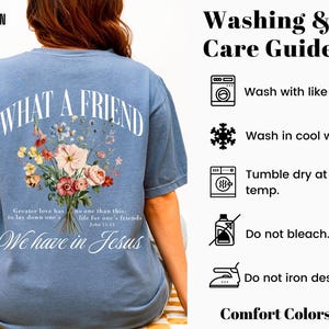 K&ouml;nnte beinhalten: Blaues Jeans-T-Shirt mit dem Text "WHAT A FRIEND" in einem Bogen. Darunter ein Blumenstrau&szlig;. Das Shirt enth&auml;lt auch den Text "We have in Jesus" und Waschanweisungen.
