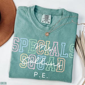May include: A light green t-shirt with the words "SPECIALS Mrs. Collins SQUAD P.E." in colourful block letters. A gold necklace with a pendant is draped across the shirt. The shirt is folded on a white surface.