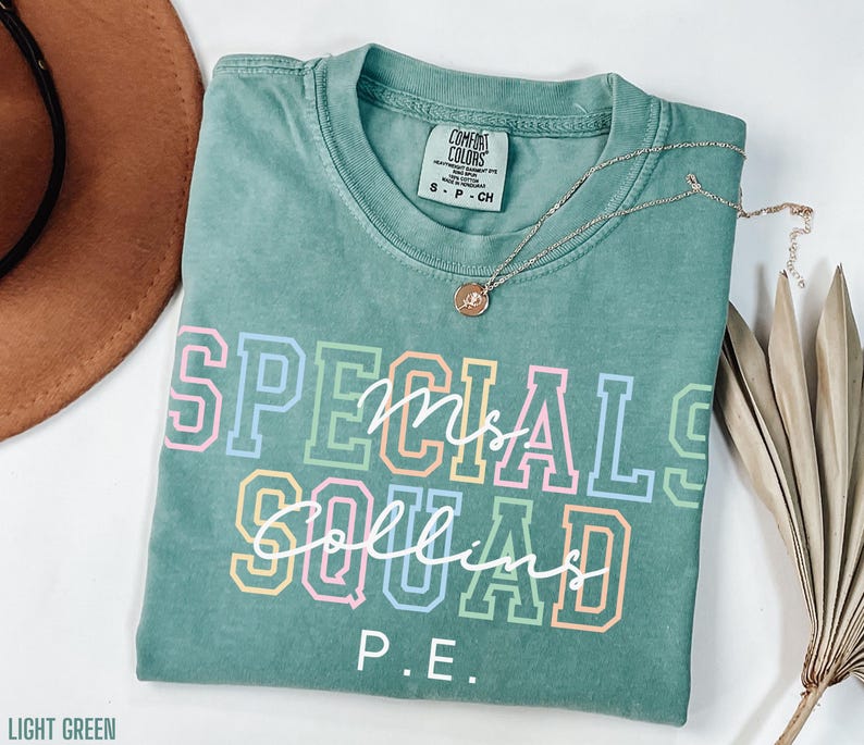 May include: A light green t-shirt with the words "SPECIALS Mrs. Collins SQUAD P.E." in colourful block letters. A gold necklace with a pendant is draped across the shirt. The shirt is folded on a white surface.