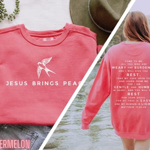May include: A pink sweatshirt with a white bird graphic and the text "JESUS BRINGS PEACE" on the front. The back of the sweatshirt has the text "COME TO ME, ALL YOU WHO ARE WEARY AND BURDENED, AND I WILL GIVE YOU REST. TAKE MY YOKE UPON YOU AND LEARN FROM ME. FOR I AM GENTLE AND HUMBLE IN HEART, AND YOU WILL FIND REST FOR YOUR SOULS. FOR MY YOKE IS EASY AND MY BURDEN IS LIGHT. MATTHEW 11:26-30"