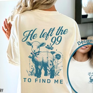 May include: Cream-coloured t-shirt with a teal graphic on the back. The design includes a lamb, flowers, and the text "He left the 99 to find me." The front design is also visible. A relaxed fit, casual style.