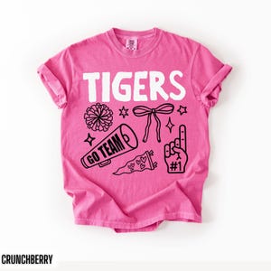 Puede incluir: Una camiseta de manga corta de color rosa brillante con el texto blanco TIGERS en la parte superior. Debajo del texto hay ilustraciones gr&aacute;ficas negras de un pomp&oacute;n, un meg&aacute;fono con las palabras GO TEAM, un lazo, un bander&iacute;n con corazones y un dedo de espuma con #1.