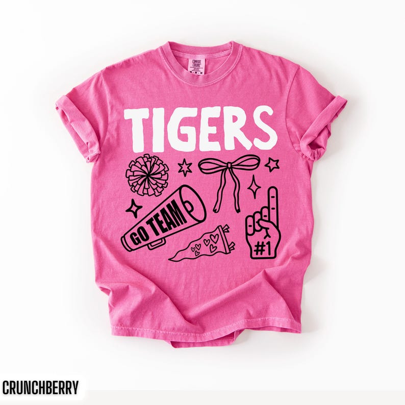 Puede incluir: Una camiseta de manga corta de color rosa brillante con el texto blanco TIGERS en la parte superior. Debajo del texto hay ilustraciones gr&aacute;ficas negras de un pomp&oacute;n, un meg&aacute;fono con las palabras GO TEAM, un lazo, un bander&iacute;n con corazones y un dedo de espuma con #1.