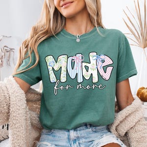 Op de afbeelding: Lichtgroen T-shirt met de tekst "MADE for more" in een kleurrijk ontwerp met bloemen- en streepmotieven. Het shirt is een casual stijl, geschikt voor dagelijks gebruik. De tekst is in een speels lettertype.