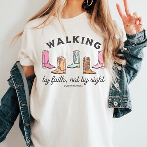 Puede incluir: Camiseta blanca con el texto "WALKING by faith, not by sight" y una fila de cinco botas de vaquero de colores. El texto "2 CORINTHIANS 5:7" est&aacute; debajo de las botas.