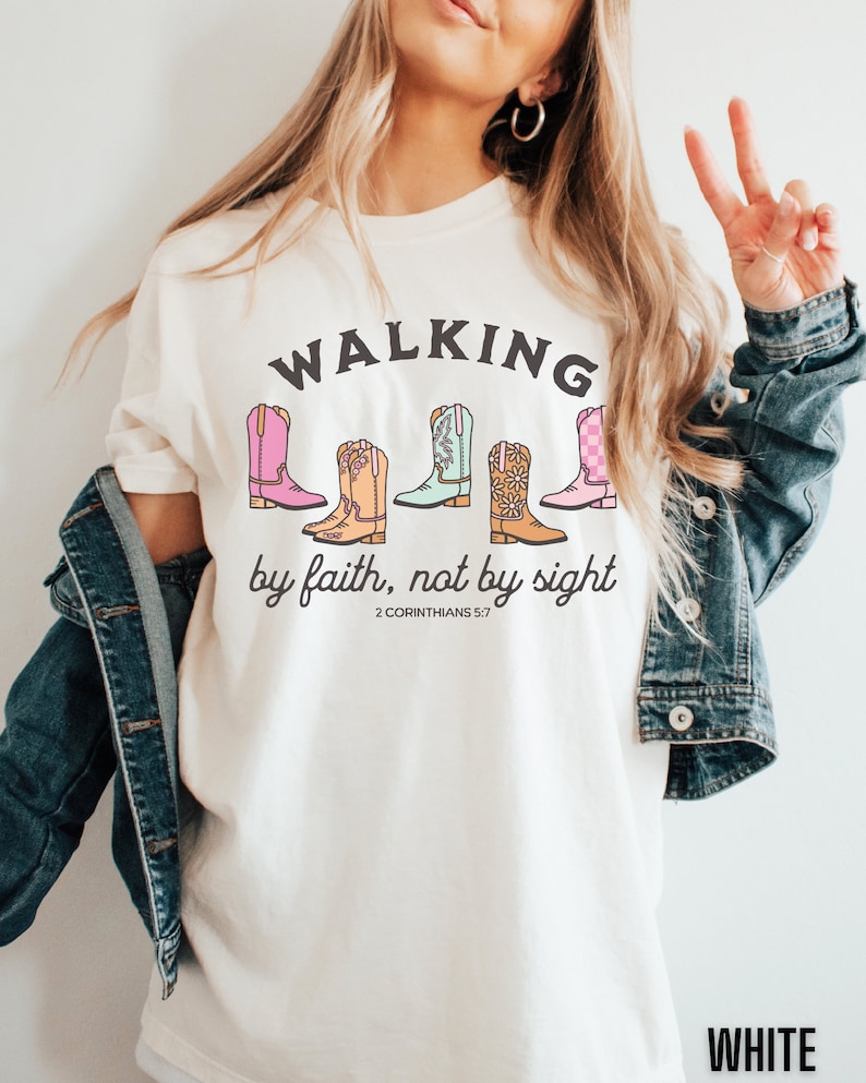 Puede incluir: Camiseta blanca con el texto "WALKING by faith, not by sight" y una fila de cinco botas de vaquero de colores. El texto "2 CORINTHIANS 5:7" est&aacute; debajo de las botas.