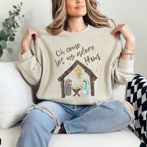 Puede incluir: Una sudadera beige con el texto "Oh come let us adore Him" y una ilustraci&oacute;n simple de la escena de la Natividad con una estrella sobre el pesebre.