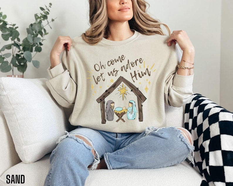Puede incluir: Una sudadera beige con el texto "Oh come let us adore Him" y una ilustraci&oacute;n simple de la escena de la Natividad con una estrella sobre el pesebre.