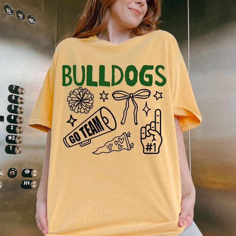 Puede incluir: Una camiseta de manga corta de color amarillo claro con el texto verde BULLDOGS y gr&aacute;ficos de l&iacute;neas negras que incluyen un pomp&oacute;n, un lazo, un meg&aacute;fono con el texto GO TEAM, un bander&iacute;n con corazones y un dedo de espuma con el texto #1.