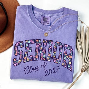 May include: A violet short-sleeved t-shirt with a crew neckline. The front features the word SENIOR in large block letters filled with a colorful floral pattern, and the text Class of 2027 written in a dark purple script font below the word.