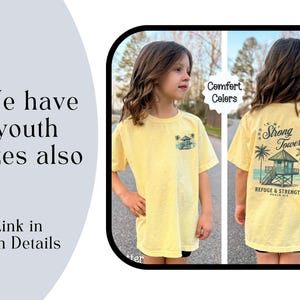 Puede incluir: Una camiseta amarilla con un gr&aacute;fico de una casa de playa y palmeras. La parte posterior de la camiseta dice "Dios es mi Torre Fuerte" y "Refugio y Fuerza Salmo 91:2". La camiseta est&aacute; disponible en tallas para ni&ntilde;os.