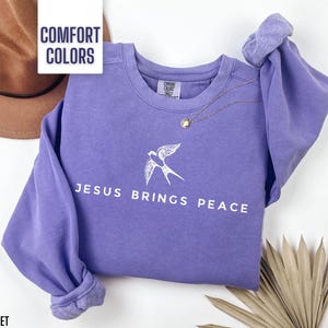 May include: A purple sweatshirt with the text "Jesus Brings Peace" and a white bird design. The sweatshirt is made of a soft, comfortable fabric.