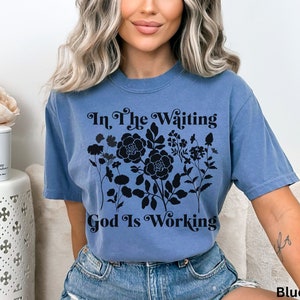 Op de afbeelding: Een jeansblauw T-shirt met zwarte tekst die "In The Waiting God Is Working" luidt en een bloemenmotief.