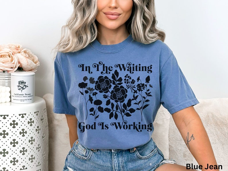 Op de afbeelding: Een jeansblauw T-shirt met zwarte tekst die "In The Waiting God Is Working" luidt en een bloemenmotief.