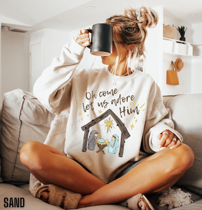 Puede incluir: Una sudadera blanca con un gr&aacute;fico de la natividad navide&ntilde;a y el texto "Oh come let us adore Him".