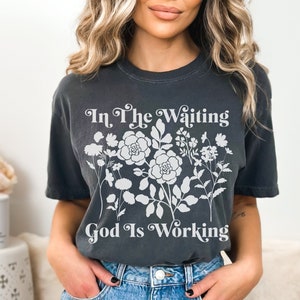 Op de afbeelding: Een antracietkleurig T-shirt met een wit bloemenmotief en de tekst "In The Waiting God Is Working".