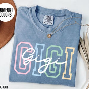 May include: A blue jean t-shirt with the word "Gigi" in colorful block letters. The shirt is made of a soft, comfortable fabric and has a vintage wash.