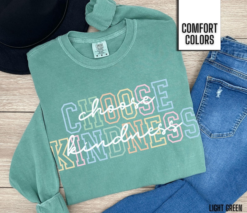 May include: A light green long-sleeved t-shirt with the text "Choose Kindness" in rainbow colors. The shirt is made of a soft, comfortable fabric and has a relaxed fit.