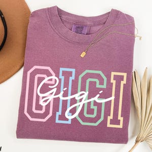May include: A mauve colored t-shirt with the word "Gigi" written in white script across the front. The word "Gigi" is outlined in rainbow colors.