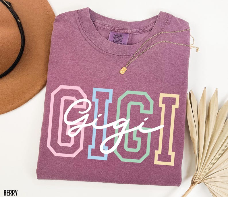 May include: A mauve colored t-shirt with the word "Gigi" written in white script across the front. The word "Gigi" is outlined in rainbow colors.